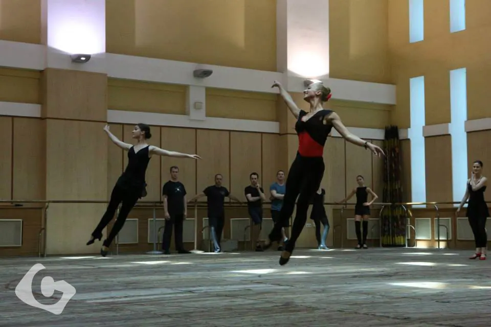 Dancers leaping mid-air during rehearsal in a studio, with other dancers watching from the barre